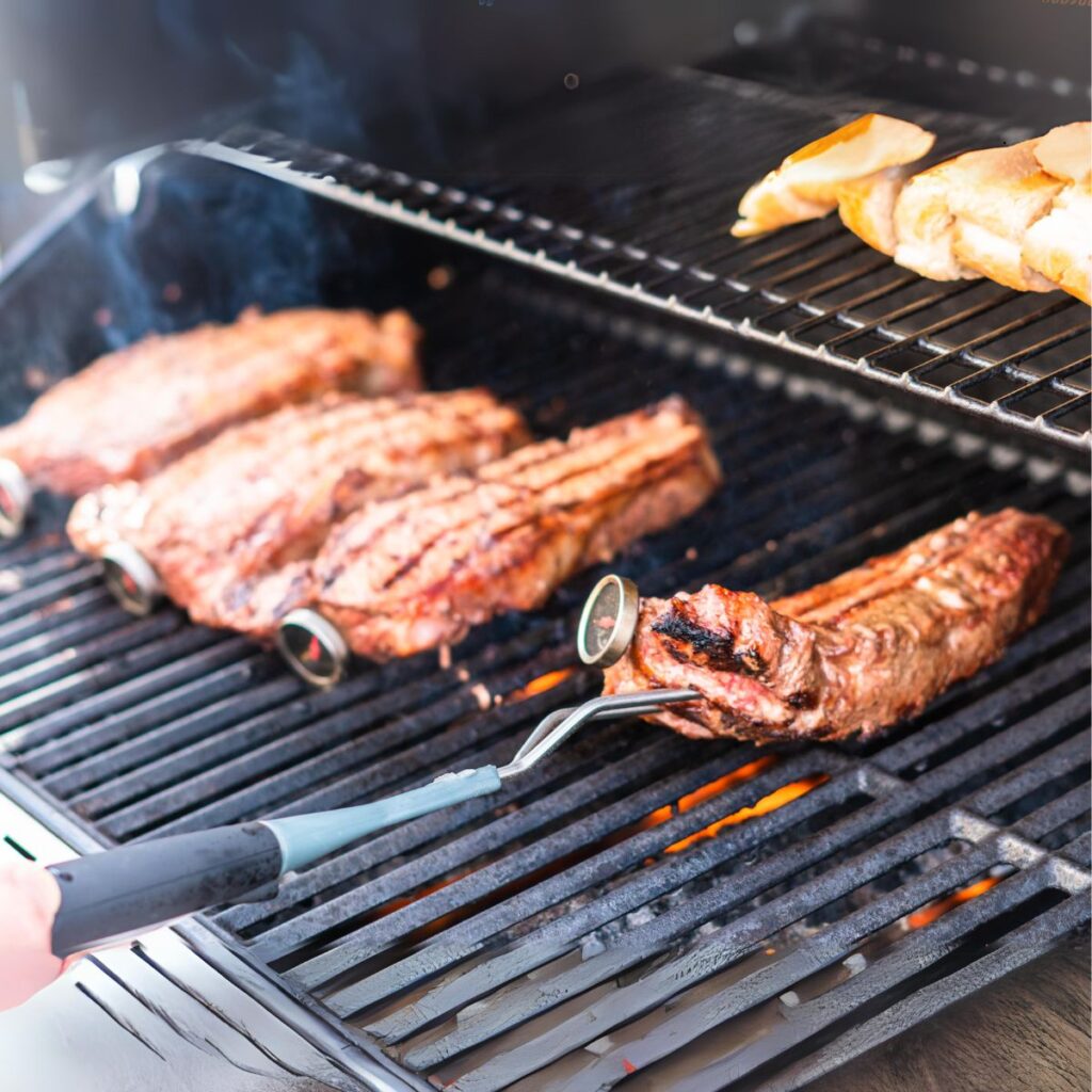 Someone with a food thong moving the New York strip steak cuts on the right side of the grill.