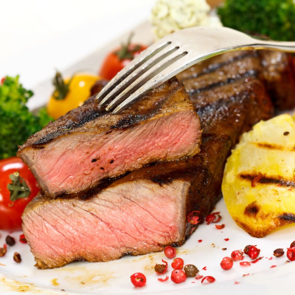A sliced New York strip steak on top of another with vegetables and spices surrounding them on a plate.