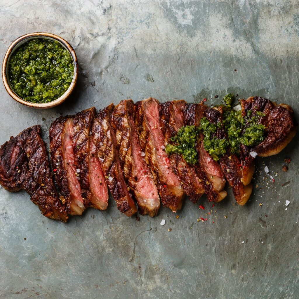 A sliced, grilled New York strip steak with chimichurri on the half top of the steak and a small bowl containing the chimichurri sauce.