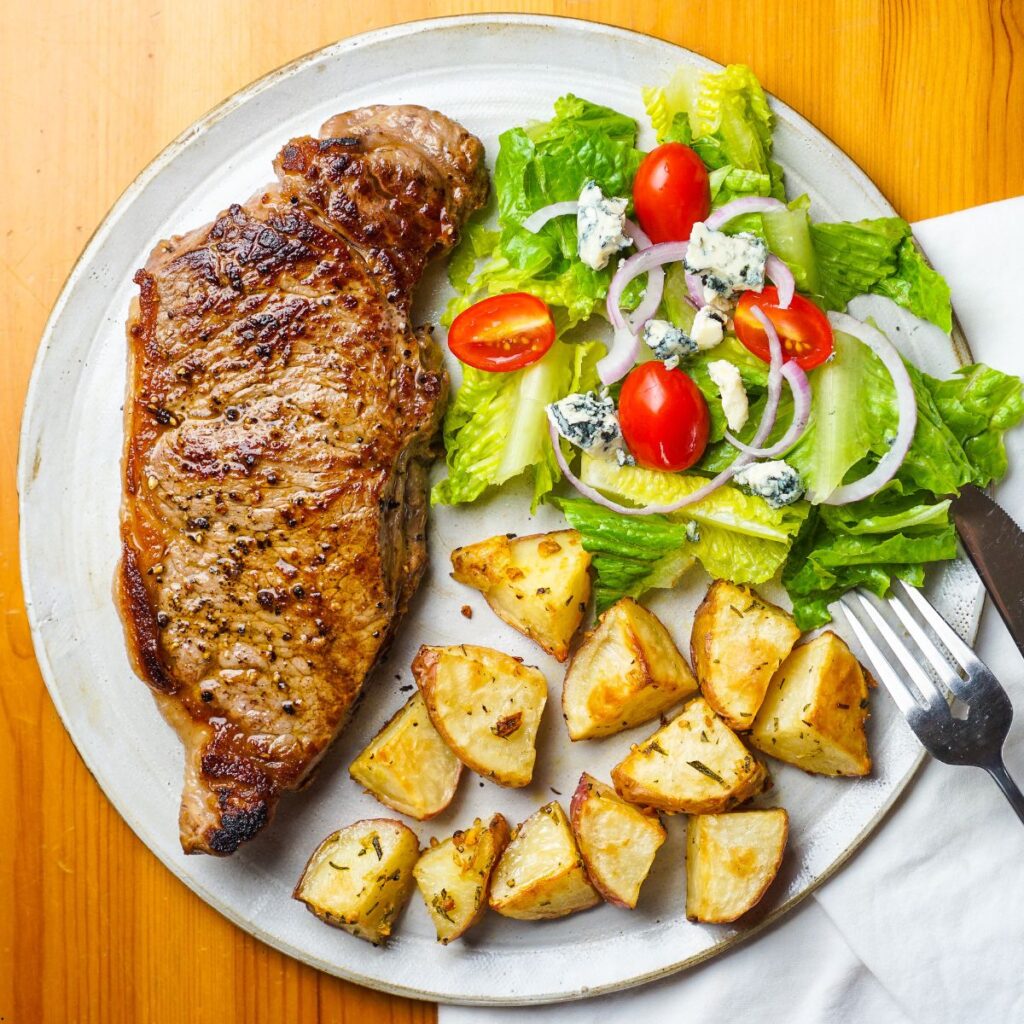 A plate of instant pot New York strip steak with fresh vegetables and roasted potatoes as side dishes.