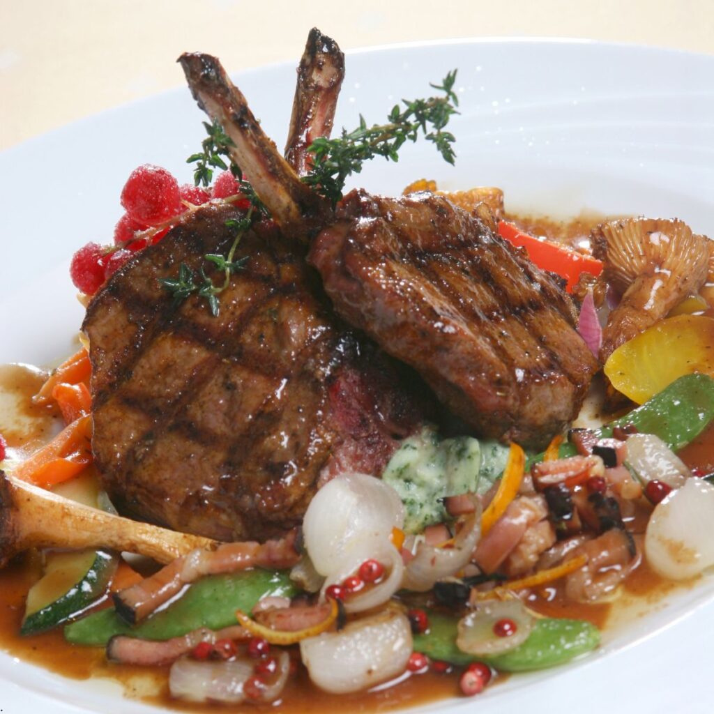 Two grilled venison chops surrounded by vegetables in sauce on a white plate.